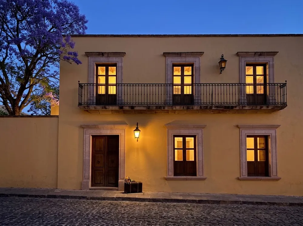 La fachada de la casa al anochecer, con todas las ventanas iluminadas, la lámpara encendida junto a la puerta y una maleta lista en la banqueta